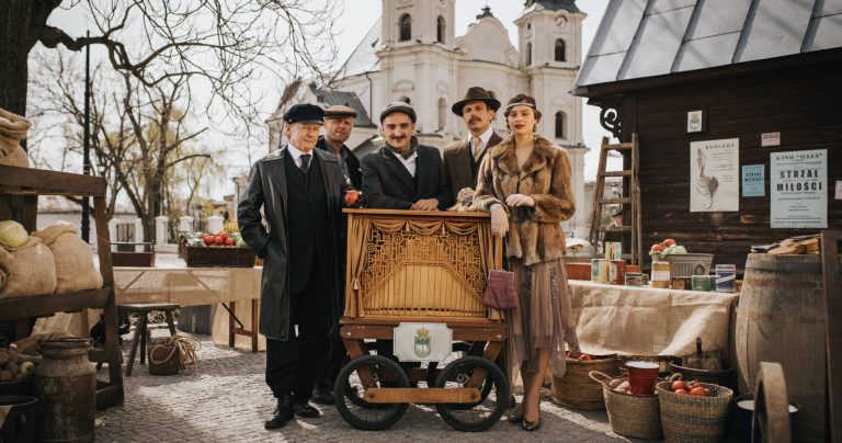 A group of people in retro costumes at a stylized stall, with the Church of the Propagation of the Apostles and the historic kiosk from Luczkowski Square in the background.   