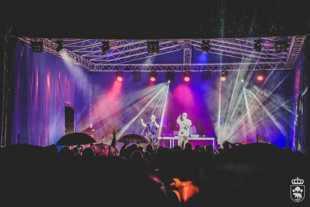 Koncert plenerowy na oświetlonej scenie podczas deszczu, z publicznością trzymającą parasole.