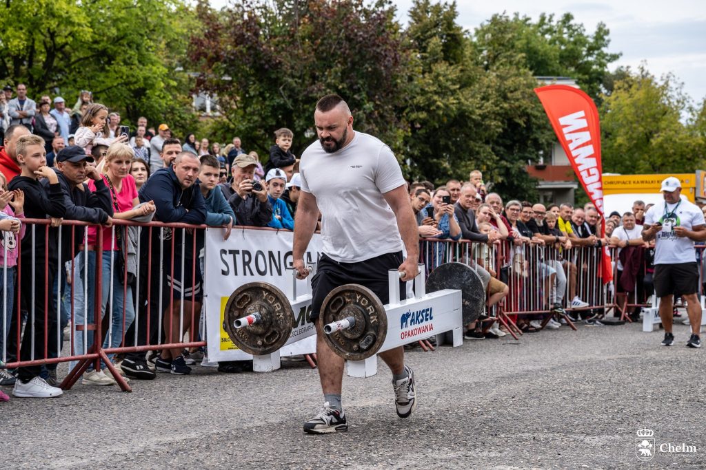 Zawodnik strongman niesie ciężary w konkurencji siłowej przed publicznością.