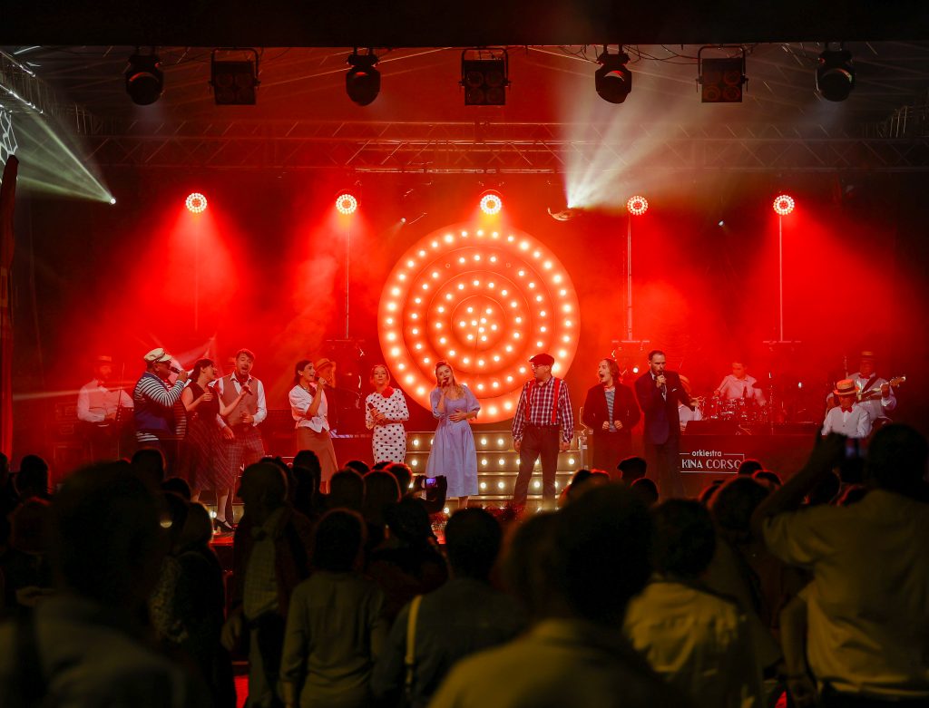 Grupa wokalistów i muzyków występuje na dużej scenie.