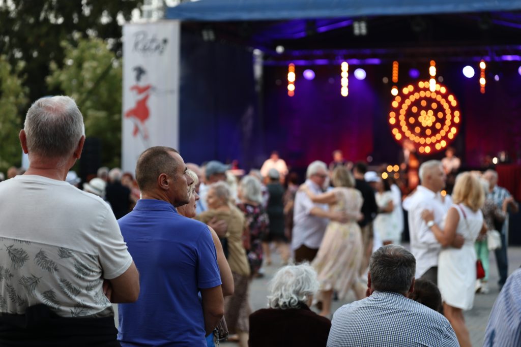 Uczestnicy wydarzenia stoją i siedzą przed sceną plenerową, na której odbywa się koncert.