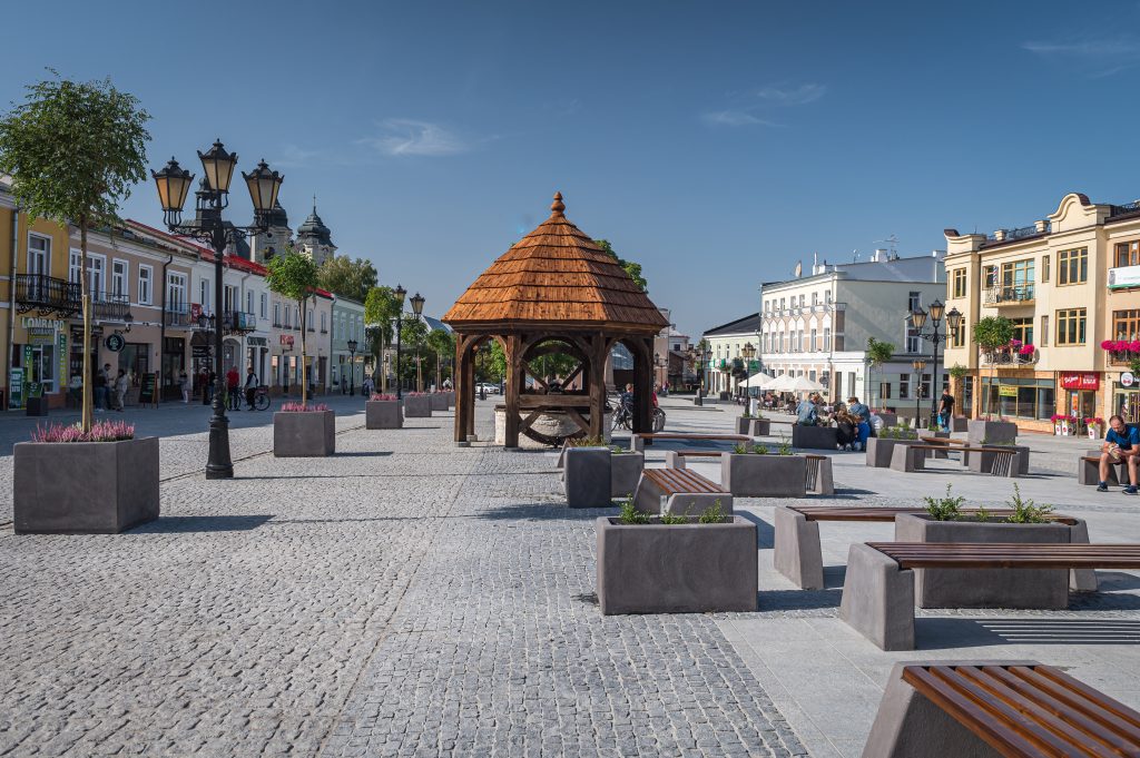 Plac Łuczkowskiego w Chełmie z zabytkową studnią, miejską zabudową i dachem kościoła pw. Rozesłania św. Apostołów w tle.